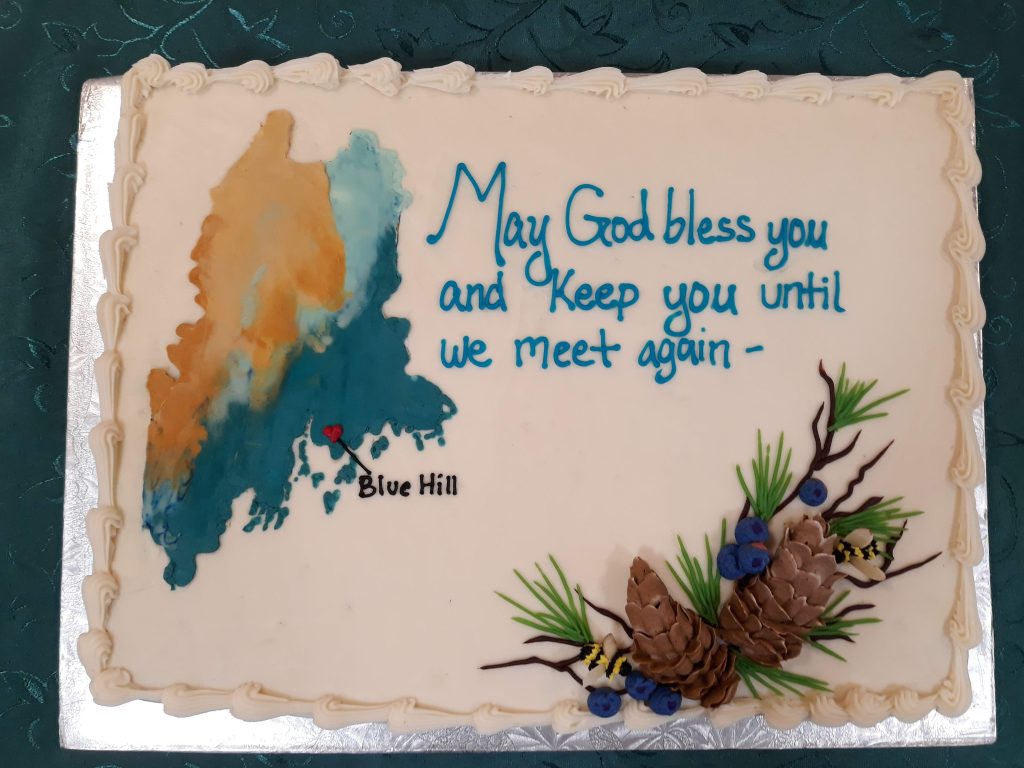 A sheet cake with a map of the state of Maine with the town of Blue Hill marked on it. The words "May God bless you and keep you until we meet again" are written in icing above a decoration with bees, huckleberries, and pine cones formed from icing.